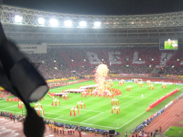 Ein Stadion mit Flutlicht, Eisengittern, Zuschauern, stehenden Individuen, Werbetafeln, Anzeigetafeln, Sicherheitskameras und dekorativen Fahnen.