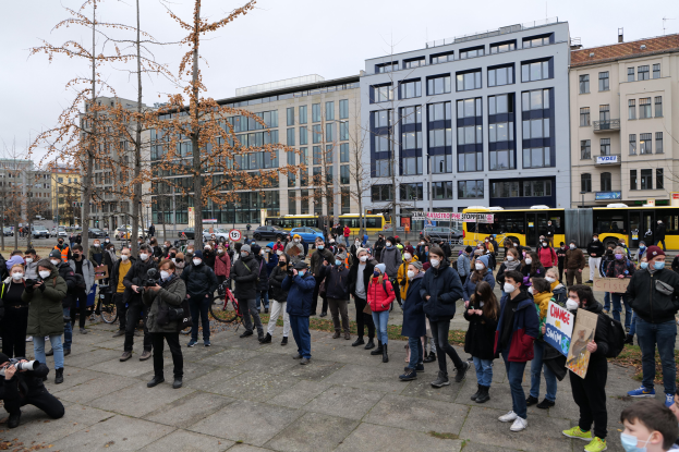 Eine große Gruppe von Menschen, einige mit Masken und Schildern, steht vor einem Gebäude während einer Protestaktion in Berlin. Im Hintergrund sind Fahrzeuge, Bäume, Laternenpfähle und Gebäude mit Fenstern sowie der Himmel zu sehen.