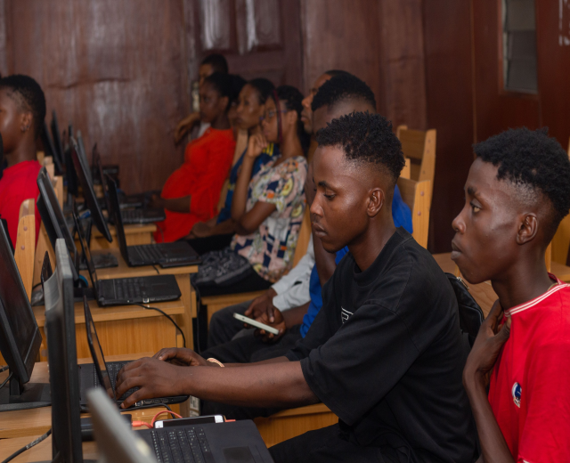 Eine Gruppe von Menschen sitzt an Schreibtischen mit Computern, Laptops und Monitoren in einem klassenzimmerähnlichen Setting mit einem Fenster im Hintergrund.