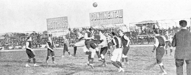 Ein Schwarz-Weiß-Bild einer Gruppe von Männern, die auf einem Feld Fußball spielen, mit einem Ball in der Luft und einer Zuschauermenge im Hintergrund, mit Schildern, die Text über das Spiel anzeigen.