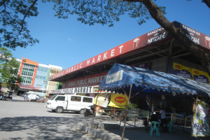 Ein belebtes Stadtmarkt-Szenario mit Gebäuden, Bäumen, Fahrzeugen, Menschen, Sitzgelegenheiten, Ständen, Bannern und einem klaren blauen Himmel.