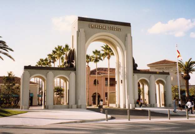 Eingang zu Universal Studios Hollywood mit einem Bogen, einer Statue, Menschen, Bäumen, Gebäuden, einem Fahnenmast mit einer Fahne und einem bewölkten Himmel.