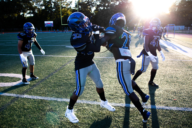 Eine Gruppe junger Männer, die Fußball auf einem von Bäumen umgebenen Feld unter einem klaren blauen Himmel spielen, alle tragen Helme und Schuhe.