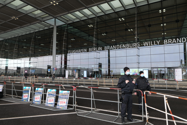 Zwei Polizisten in schwarzen Uniformen stehen vor einem großen Glasgebäude mit Text darauf, mit Absperrungen und Texttafeln im Vordergrund und anderen Menschen, Pfählen und Lichtern im Hintergrund.