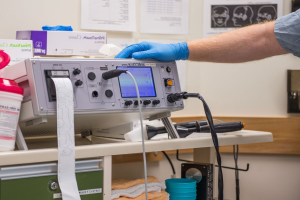 Eine Person in blauen Handschuhen arbeitet an einem medizinischen Gerät auf einem Tisch, der mit verschiedenen Gegenständen übersät ist, während im Hintergrund Papiere an der Wand hängen und damit eine Krankenhausatmosphäre suggerieren.