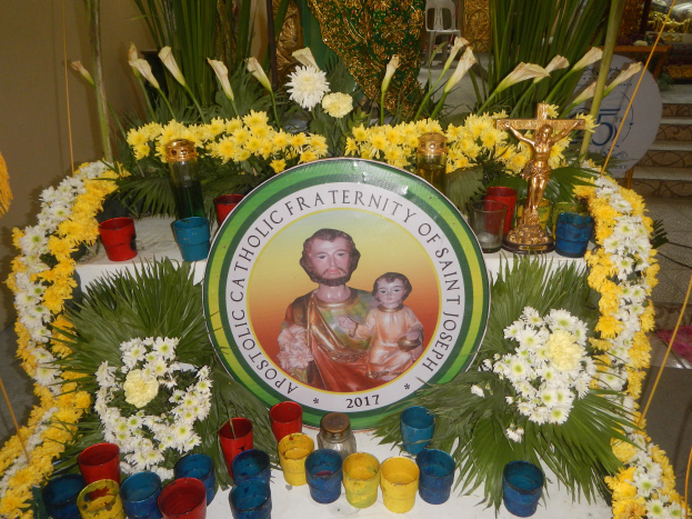 Ein mit bunten Blumen, Kerzen und einer Tafel mit einem Porträt und Text geschmückter Tisch, vor einer Kulisse aus Pflanzen, Stühlen und anderen Gegenständen, schafft eine festliche Atmosphäre für die Bruderschaft von St. Joseph.