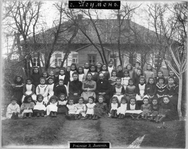 Schwarzes und weißes Foto einer Gruppe von Kindern, die vor einem Gebäude mit Fenstern posieren, umgeben von Bäumen und einem klaren Himmel, mit Text unten, der "Jugoslawien, die erste Schule der Welt" lautet.
