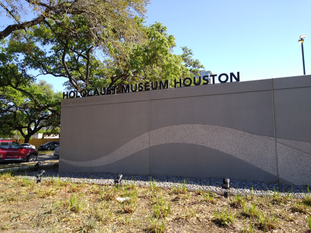 Außenansicht des Houston Holocaust Museums mit einer Gedenkwand, Steinen, Grünflächen, Bäumen, vorbeifahrenden Fahrzeugen, einem Straßenschild und einem bewölkten Himmel.