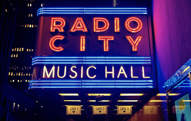 Hell erleuchtetes Radio City Music Hall in New York City mit hohen umliegenden Gebäuden, das eine 'Radio City'-Leuchtschrift und mehrere kleinere Schilder auf der rechten Seite zeigt.