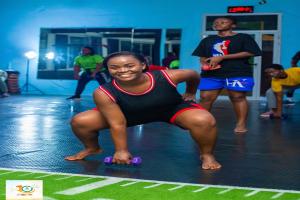 Eine Frau in einem Fitnessstudio führt eine Kniebeuge mit Hanteln aus, wobei verschiedene Geräte und Logos im Hintergrund zu sehen sind.