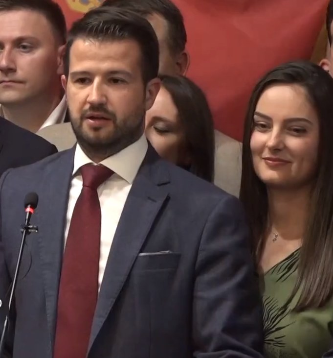 Ein Mann im Anzug spricht an einem Rednerpult mit einem Mikrofon, während er eine Menge hinter einem Banner anspricht.