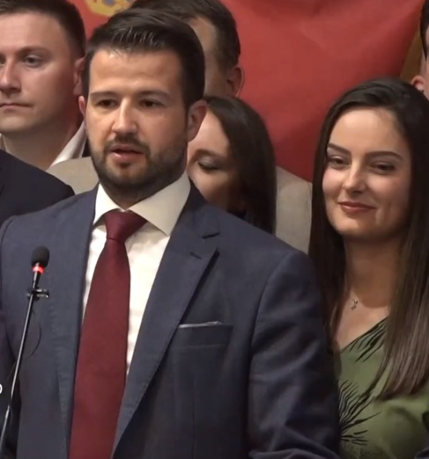 Ein Mann im Anzug spricht an einem Rednerpult mit einem Mikrofon, während er eine Menge hinter einem Banner anspricht.