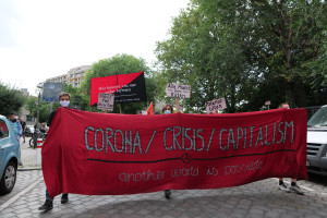 Eine Gruppe von Menschen marschiert mit einer roten Fahne, auf der 'Corona-Krise/Kapitalismus' steht, durch eine Straße. Im Hintergrund sind geparkte Fahrzeuge, Gebäude und Bäume unter einem klaren blauen Himmel zu sehen.