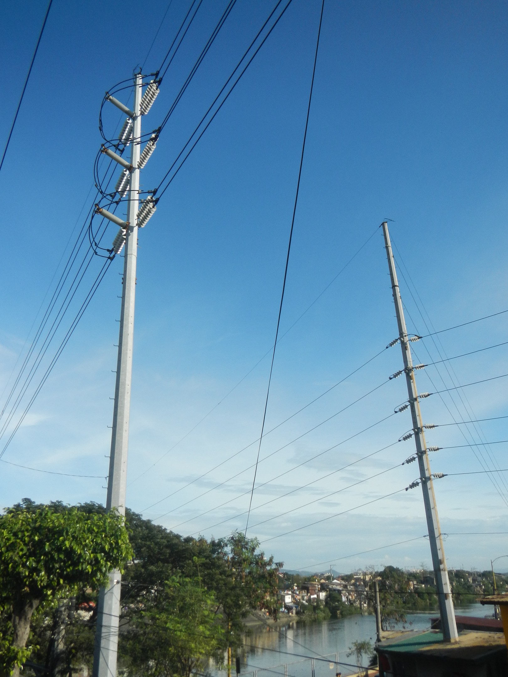 Stromleitung mit Strommasten und Drähten, die neben einer Straße an einem Gewässer entlangführen, mit Bäumen und Gebäuden im Hintergrund und einem klaren blauen Himmel.