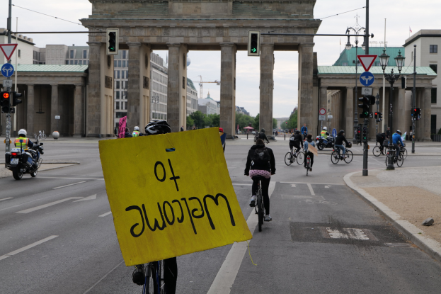 Eine Gruppe von Menschen in Helmen fährt mit Fahrrädern eine Straße entlang vor dem Brandenburger Tor in Berlin, Deutschland, wobei eine Person ein gelbes Schild hält, Lichtmasten, Verkehrszeichen, Gebäude, Bäume und einen klaren blauen Himmel im Hintergrund zu sehen sind.