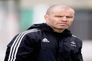 Ein kahlköpfiger Trainer in einer schwarzen Jacke steht auf einem Fußballfeld, trägt eine marineblaue Jacke mit weißen Streifen und hat die Hände mit ernster Miene verschränkt.