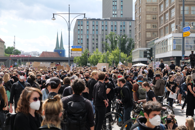 Große Gruppe von Menschen mit Masken, die auf einer von Bäumen gesäumten Straße mit Fahrrädern, Gebäuden und Ampeln in Berlin bei einer Demonstration gehen.
