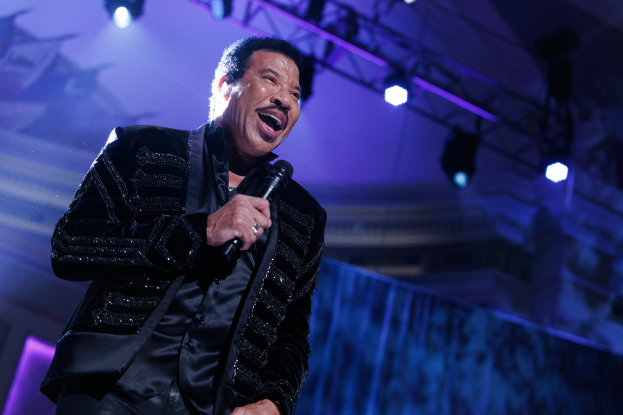 Lionel Richie performs bei den Grammys in einem schwarzen Kleid und hält ein Mikrofon in der Hand, während die Bühne von hellen Lichtern erleuchtet wird.