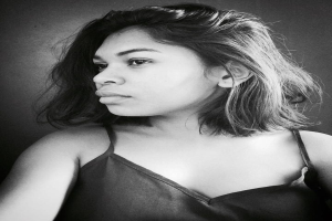 Eine selbstbewusste Frau in einem schwarzen Kleid posiert in einem Schwarz-Weiß-Foto, ihr glatter Bob und das dezente Make-up unterstreichen ihre selbstbewusste Haltung vor einer Wand.