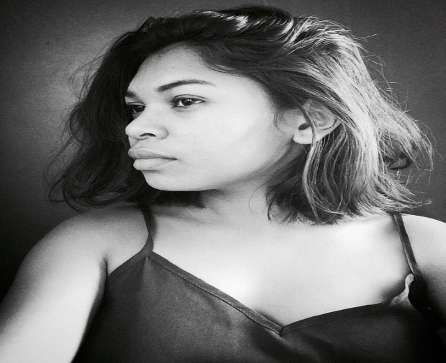 Eine selbstbewusste Frau in einem schwarzen Kleid posiert in einem Schwarz-Weiß-Foto, ihr glatter Bob und das dezente Make-up unterstreichen ihre selbstbewusste Haltung vor einer Wand.