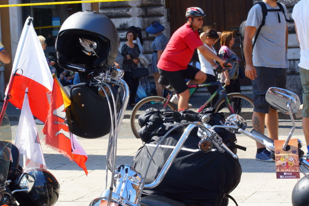 Mehrere Motorräder mit Helmen und Fahnen sowie eine schwarze Tasche und Personen im Hintergrund, eine sitzend auf einem Fahrrad und mit Sonnenbrille sowie Helm.