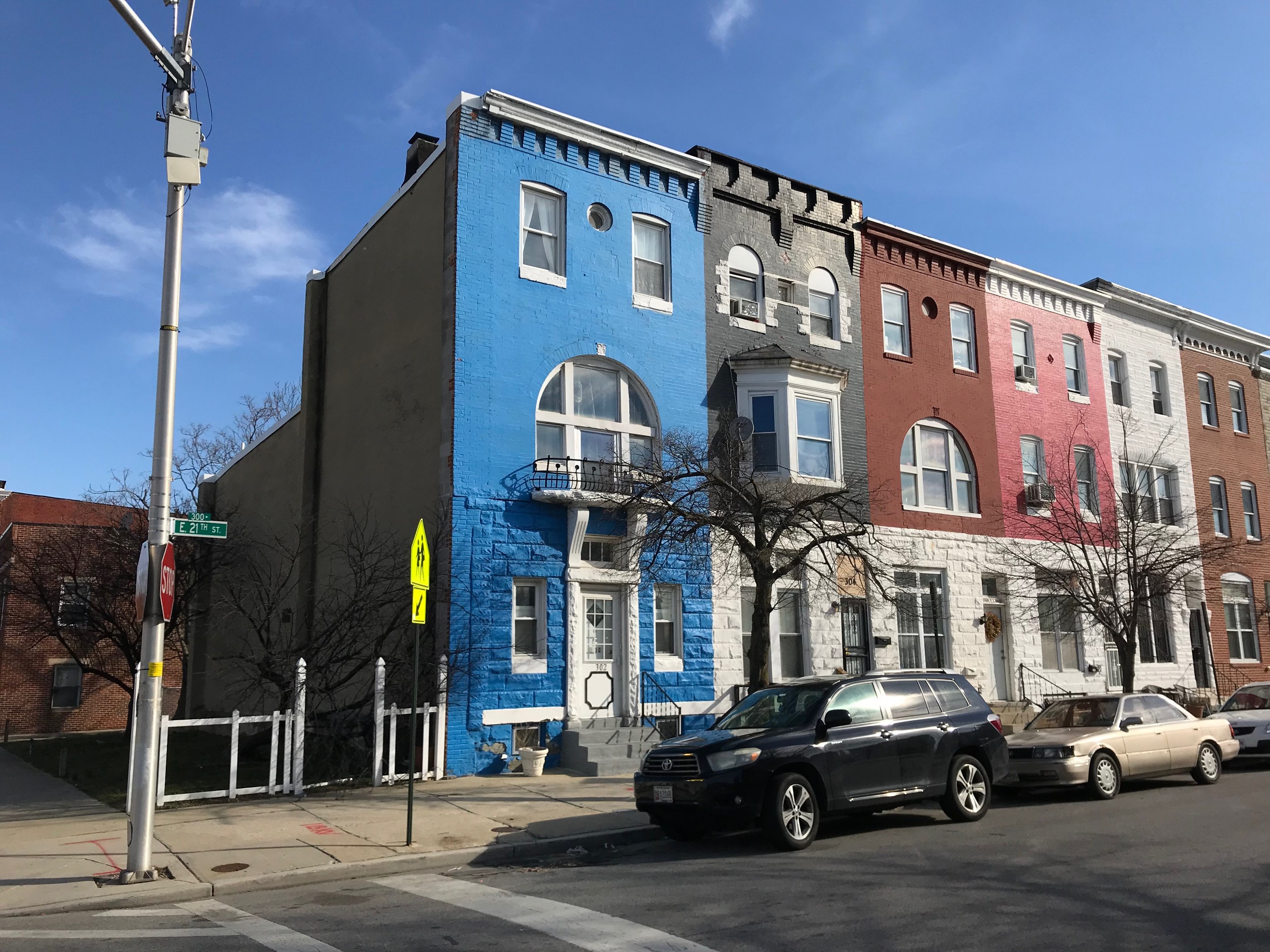 Eine Reihe farbenfroher Häuser an einer Straßenecke in Baltimore, Maryland, mit geparkten Autos, Bäumen, Pfählen, Brettern, einem Zaun und einem bewölkten Himmel.