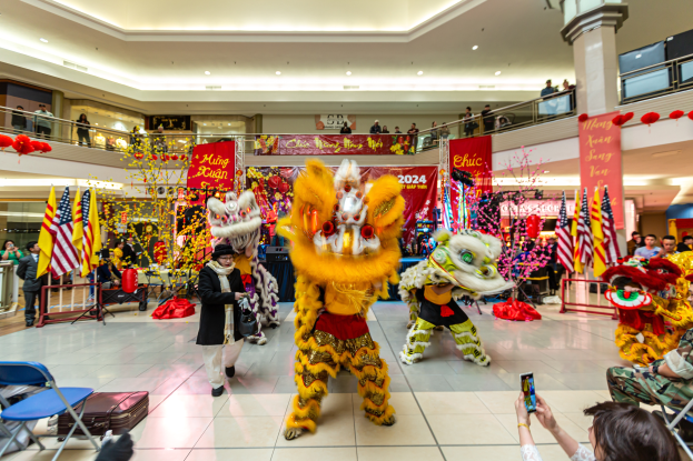 Ein lebendiger Chinese New Year-Feier in einem Einkaufszentrum mit tanzenden Menschen in Kostümen und anderen, die auf Stühlen sitzen und mobile Telefone halten, umgeben von Fahnen, Bannern, Dekorationen und Lichtern.