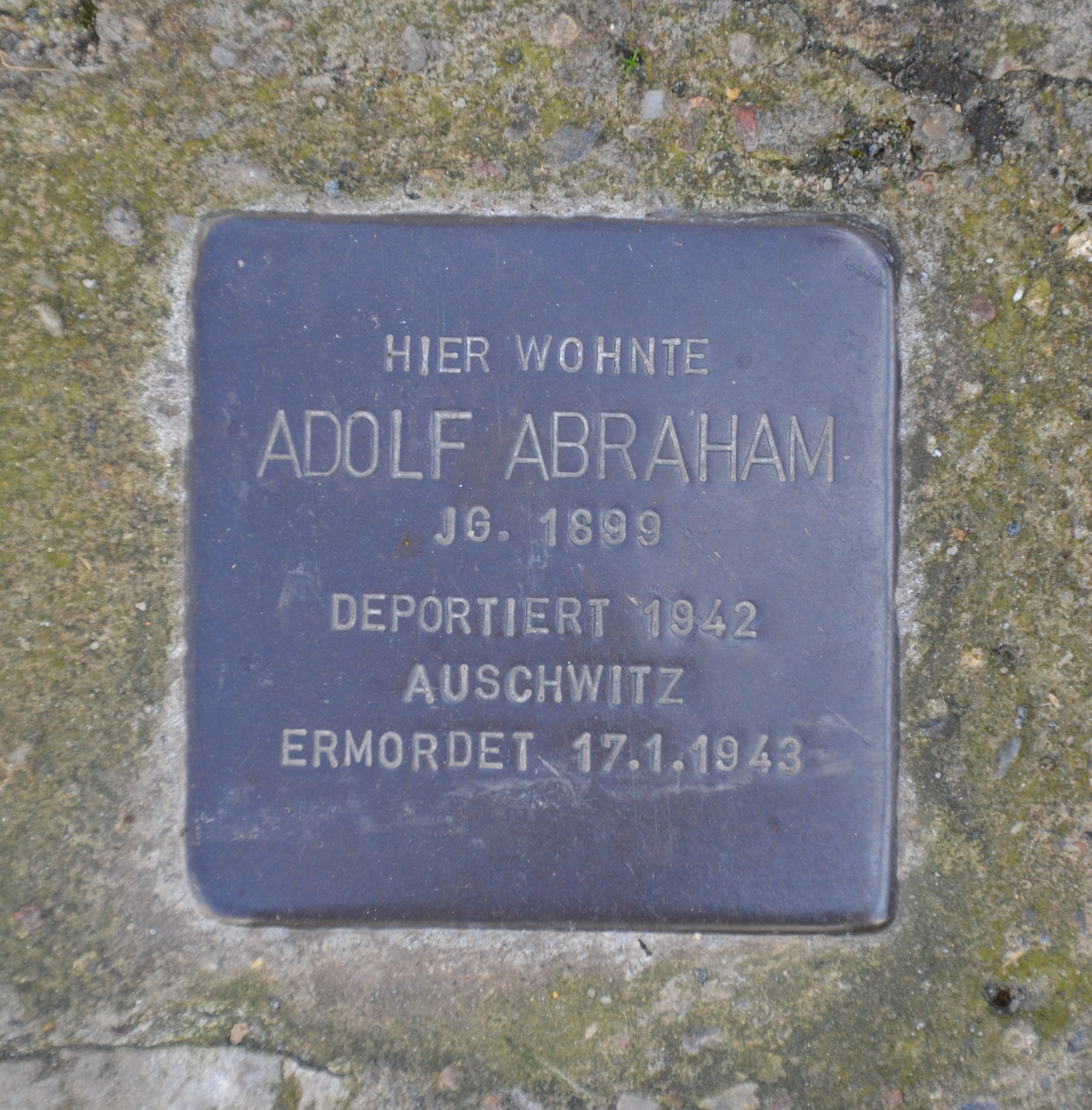 Ein rechteckiges Schild mit der Inschrift "Adolf Abraham" auf einer Steinwand angebracht.