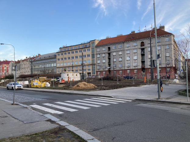 Eine Stadtstraße mit parkenden Autos, Laternen, Bäumen und Gebäuden unter einem bewölkten Himmel, mit einer Baustelle für ein neues Wohngebäude im Vordergrund.