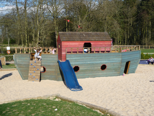 Ein Piratenschiff-Spielhaus mit einer Rutsche in der Mitte, umgeben von Sand und Gras, mit mehreren Menschen darauf spielend und Bäumen und einem klaren blauen Himmel im Hintergrund.