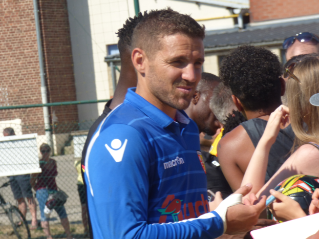 Ein Mann in einem blauen Hemd signiert Autogramme für eine Gruppe von Menschen, einige tragen Kappen und Schutzbrillen, mit einem Gebäude, einem Maschendrahtzaun und einem Fahrrad im Hintergrund.