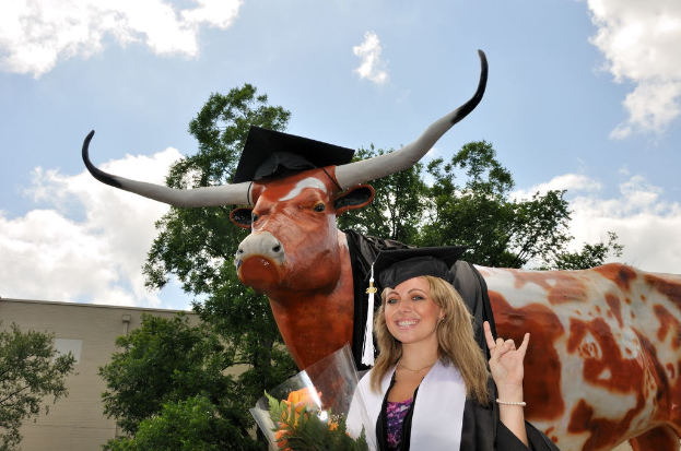 Eine Frau in einer Graduationskappe und Robe lächelt, eine Blume haltend, vor einer Skulptur, Bäumen, einem Gebäude und einem bewölkten Himmel.