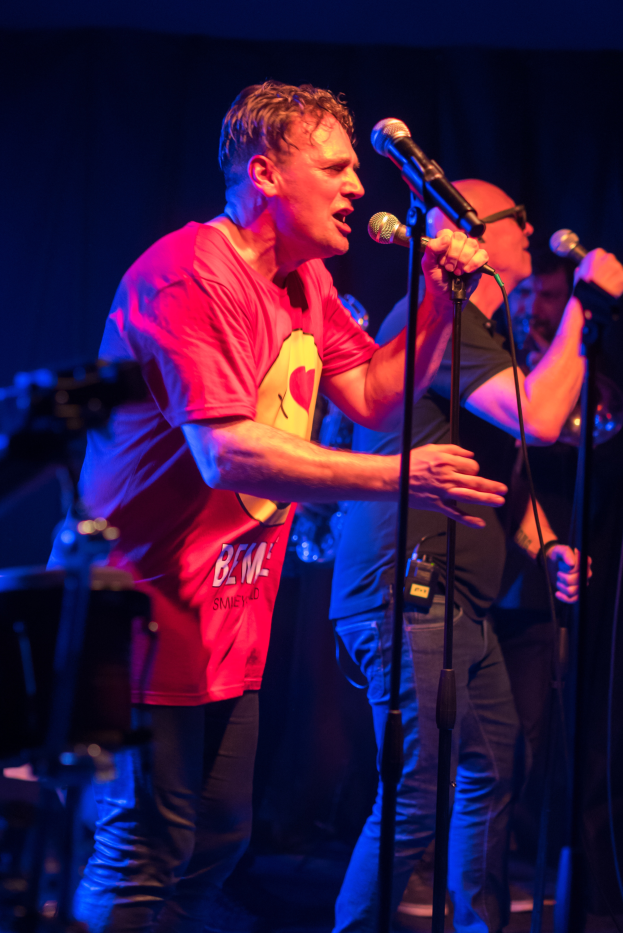 Ein Mann in rotem Hemd und Brille singt in ein Mikrofon auf der Bühne, umgeben von Musikern.