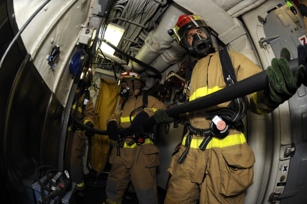 Feuerwehrleute in Helmen und Handschuhen mit einer Schlauchleitung in einem Flugzeug mit Ausrüstung und Text unten.