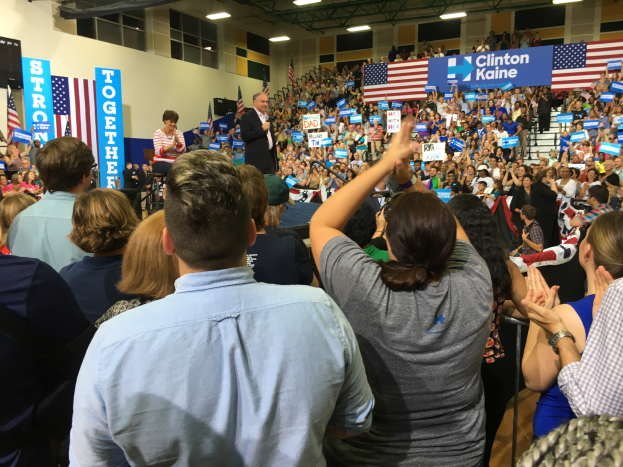 Eine große Menschenmenge klatscht und jubelt vor einer Bühne, auf der ein Mann in ein Mikrofon spricht, während eine Frau neben ihm steht; Fahnen, Banner und ein Lautsprecher sind auf dem Bühnenhintergrund zu sehen.