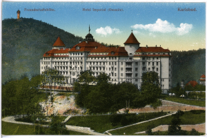 Eine alte Postkarte des Hotel Imperial in Karlsbad, Deutschland, zeigt ein großes Gebäude mit Fenstern, umgeben von Bäumen, Pflanzen, Gras, einem Pfad, einer Gruppe von Bäumen, Hügeln und einem bewölkten Himmel, mit Text oben.