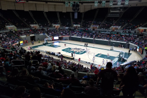 Eine große Arena voller Zuschauer bei einem Basketballspiel, mit einem Spielfeld in der Mitte und Anzeigetafeln, Scheinwerfern und anderen Gegenständen oben.
