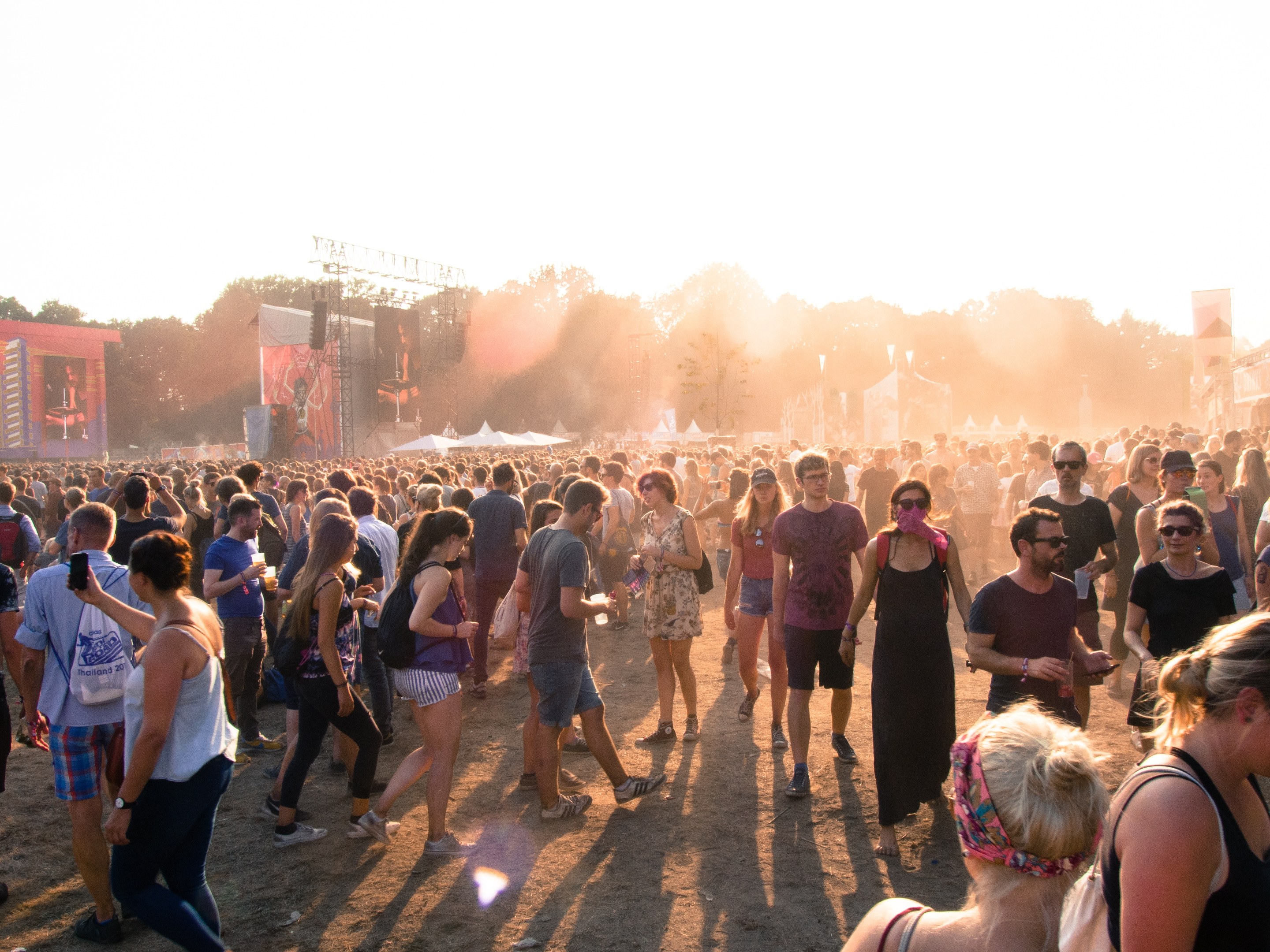 Eine große Menschenmenge, die einen Schotterweg auf einem Musikfestival entlanggeht, umgeben von Zelten, Gebäuden, Bäumen und einem klaren blauen Himmel, wobei einige Taschen tragen.