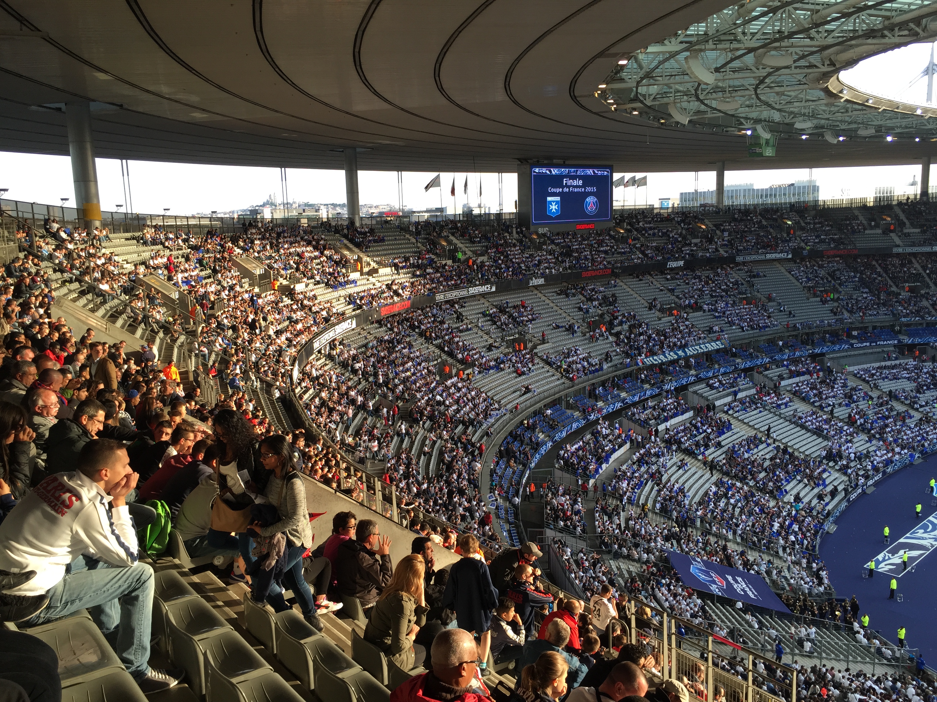 Eine große Menschenmenge sitzt im Allianz Stadion in München, Deutschland, und schaut ein Fußballspiel. Rechts ist eine Bühne mit ein paar Leuten darauf und im Hintergrund sind Fahnen, Stangen und ein Bildschirm zu sehen.