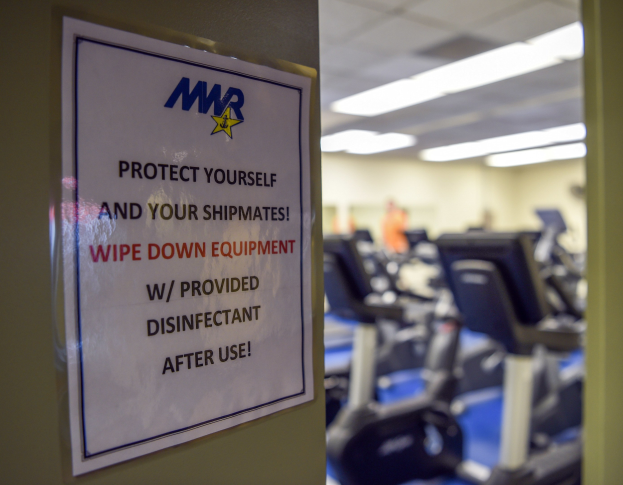 Zeichen an einer Turnhallentür mit der Aufschrift "Schützen Sie sich und Ihre Schiffskameraden" mit Laufbändern im Hintergrund und Deckenlampen.