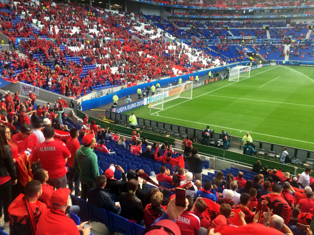 Eine große Menschenmenge in einem Stadion schaut einem Fußballspiel zu, mit Sitzplätzen und Stehplätzen, Bannern, einem Tor und Überdachungsleuchten.