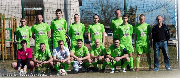 Eine Gruppe von Männern in grünen T-Shirts steht auf einem Fußballfeld mit einem Ball und einem Tor, mit Bäumen, Gebäuden und Fahrzeugen im Hintergrund.