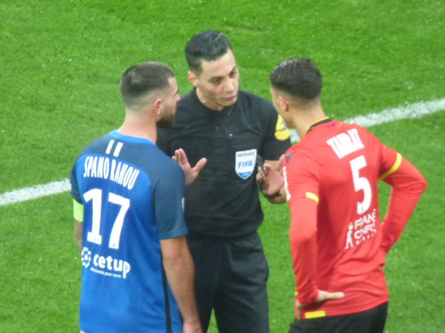 Schiedsrichter in schwarzem Hemd und Hose, der mit zwei Männern in blauen und roten T-Shirts auf einem Fußballfeld spricht, alle tragen Brillen.