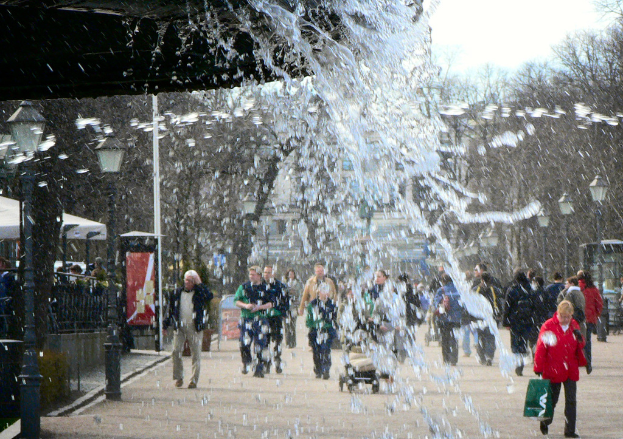 Wasser läuft aus einer unsichtbaren Quelle, mit einer Gruppe von Menschen, die auf der Straße darunter gehen, Straßenlaternen links und Bäume rechts.
