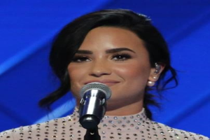 Demi Lovato steht mit einem Mikrofon auf der Bühne der Grammys, tr├Ągt ein schwarzes Kleid mit einem eleganten Bob, vor einem blauen Hintergrund, mit einem selbstbewussten Ausdruck.