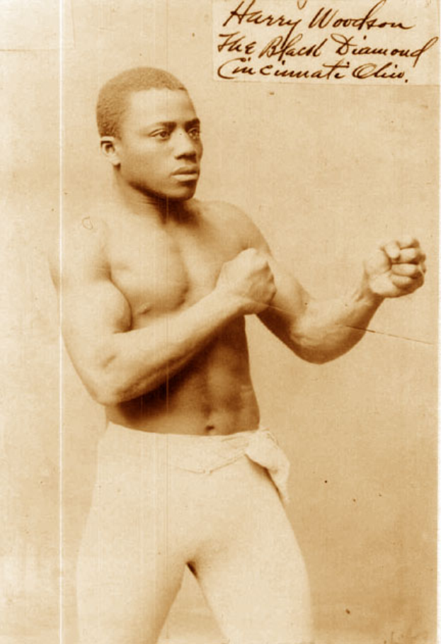 Ein altes Foto eines Mannes im Boxring, der Handschuhe und Shorts trägt und einen entschlossenen Gesichtsausdruck hat, kurze dunkle Haare und fokussierte Augen, aufrecht und stolz dasteht.