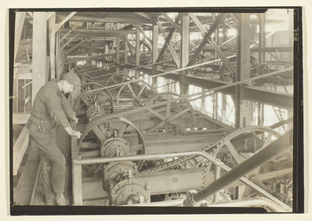 Ein Schwarz-Weiß-Foto eines Mannes, der an einer Maschine arbeitet und von Metallstangen und Rädern umgeben ist.
