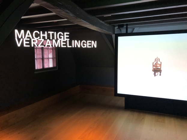 Installationsansicht der Ausstellung 'Machtige Sammlung' im Museum of Modern Art in Berlin, mit einem großen Bildschirm mit Text an der Wand dahinter und einem Fenster links.