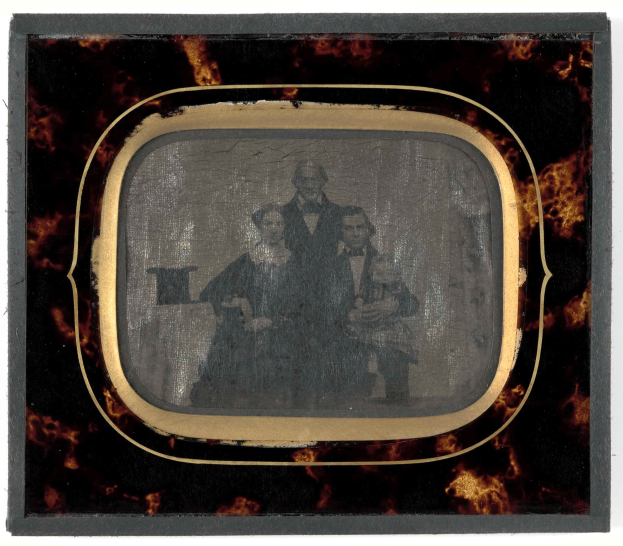 Ein altes Foto eines Mannes im Anzug und Krawatte und einer Frau mit ernstem Gesichtsausdruck vor einem Fernseher mit weißem Hintergrund.