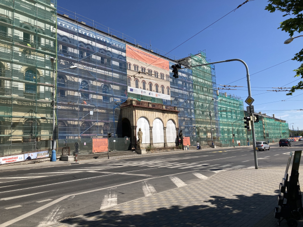 Großes modernes Gebäude im Bau in Berlin, umgeben von Straßeninfrastruktur, Fahrzeugen, Fußgängern und Bäumen gegen einen bewölkten Himmel.